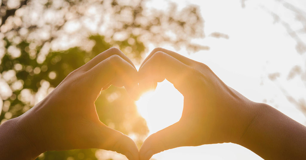 hands making heart sign against sun behind trees