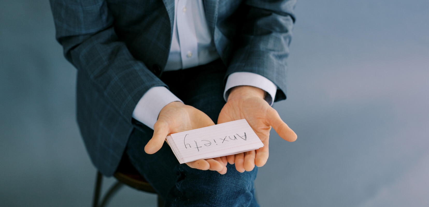 a person wearing a suit sitting in a chair holds a stack of cards, the top one reads "Anxiety"