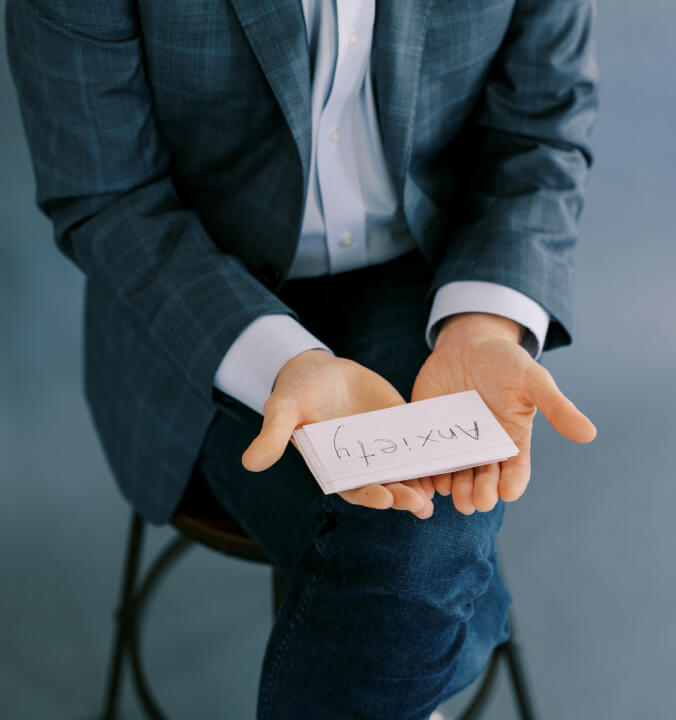 a person wearing a suit sitting in a chair holds a stack of cards, the top one reads "Anxiety"