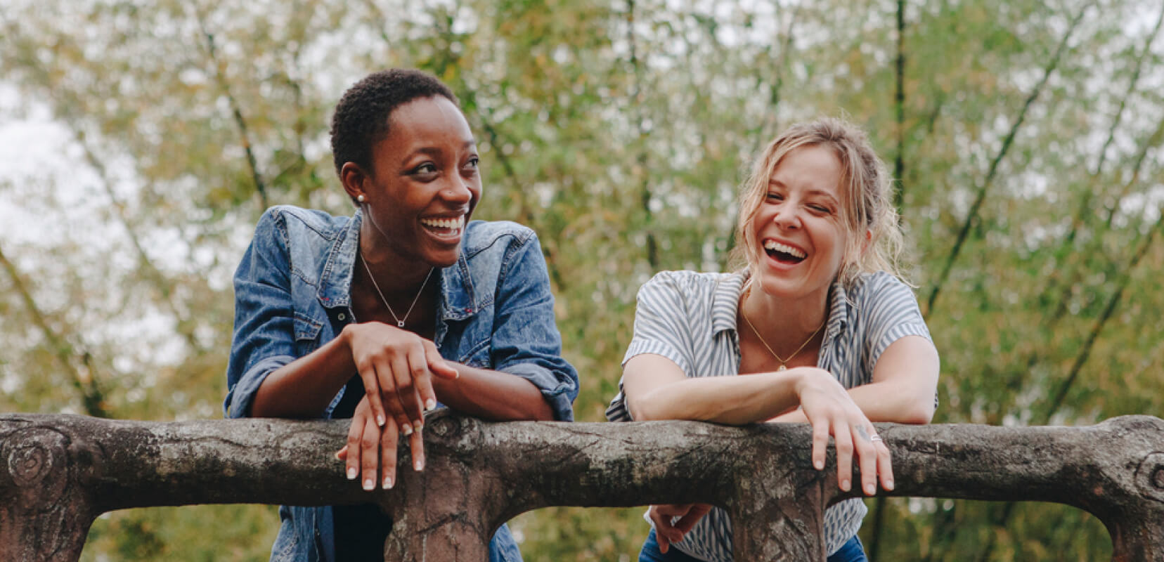Two young women, one black, one white leaning over a fence in nature, laughing