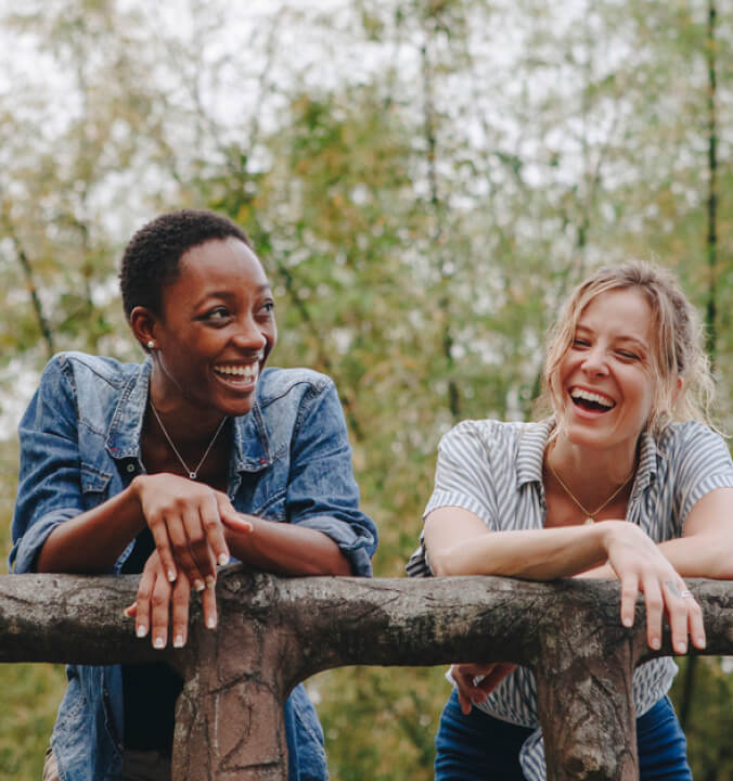 Two young women, one black, one white leaning over a fence in nature, laughing